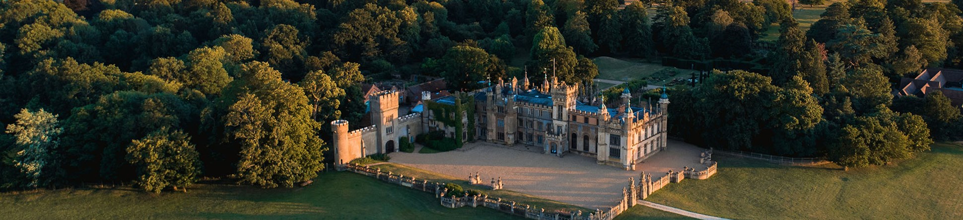 An aerial photograph shows the Knebworth House and Garden surrounded by trees with fields in the distance and a setting sunset.
