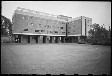 Black and white photo of three-storey building with ground floor raised on stilts leaving the ground space open. It has window ribbons and no ornamentation.