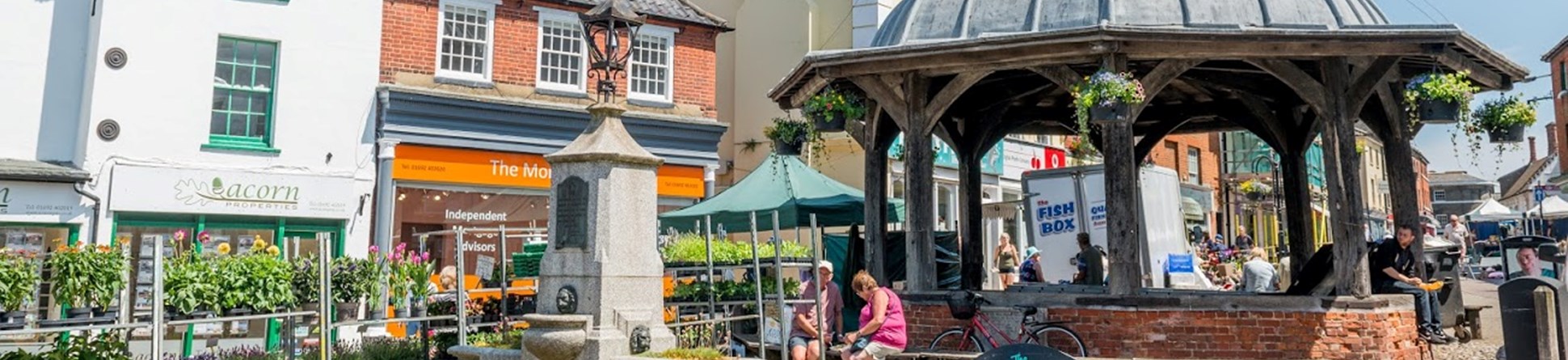 A market place with a market cross covered with a cupola, with historic buildings in use as shops in the background.