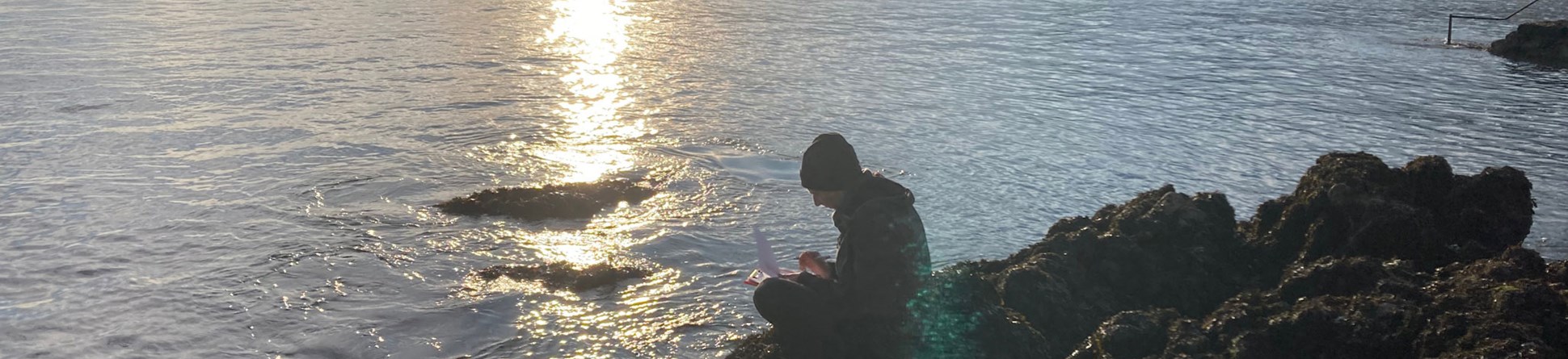 A man and a woman recording a rocky foreshore with the sea and coastal buildings in the background.