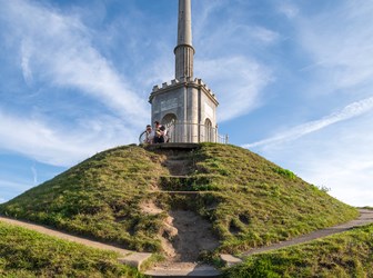 A grassy mound with a tall stone monument rises in front of a blue sky. 