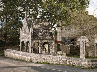 A photograph of a small, medieval stone structure enclosed by a stone wall. It has open arches around four sides. Beside it is a stone cross.