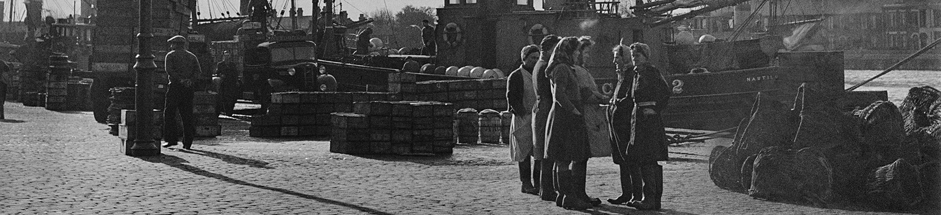 A group of women on the quay at Great Yarmouth with herring boats beyond.