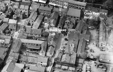 This black and white 1928 aerial photograph shows a view of part of the North Walsham Market Place, with tightly packed houses, a pub and a hotel in the distance.