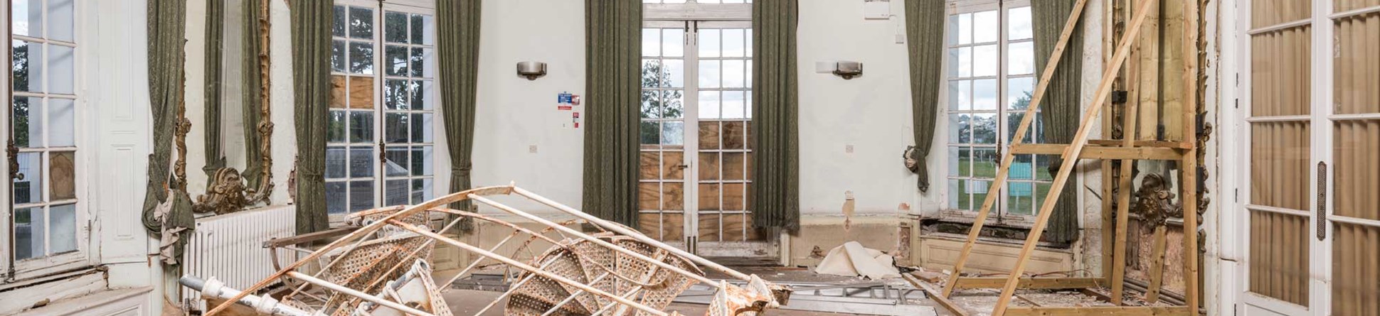 The derelict interior of a formal mansion room, with debris on the parquet flooring, a piano, and holes in the plasterwork. 