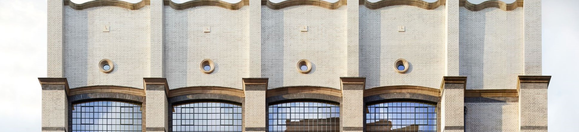 Front view of a white brick building with a wavy roof, large windows and portholes