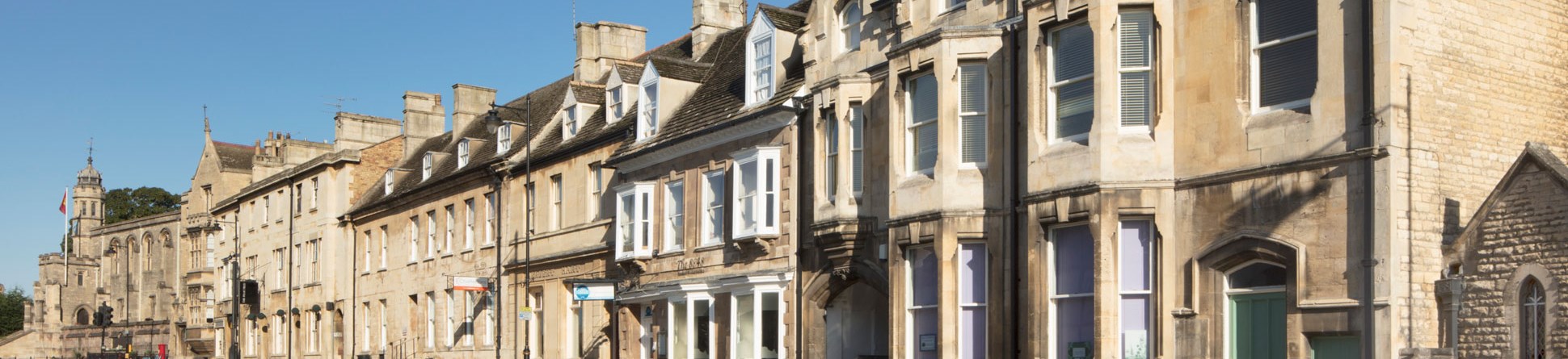 Colour photo showing a view along a row of three-storey 19th century buildings built in a light ashlar stone. Parking bays line the road in front of the buildings. Two cars are parked out front.