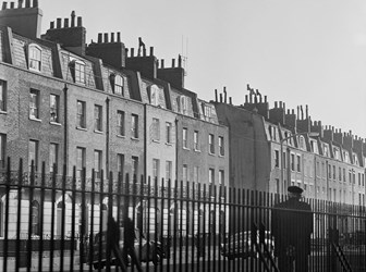 A black and white photograph of rows of tall terraced houses viewed through railings with a man walking past in the foreground.