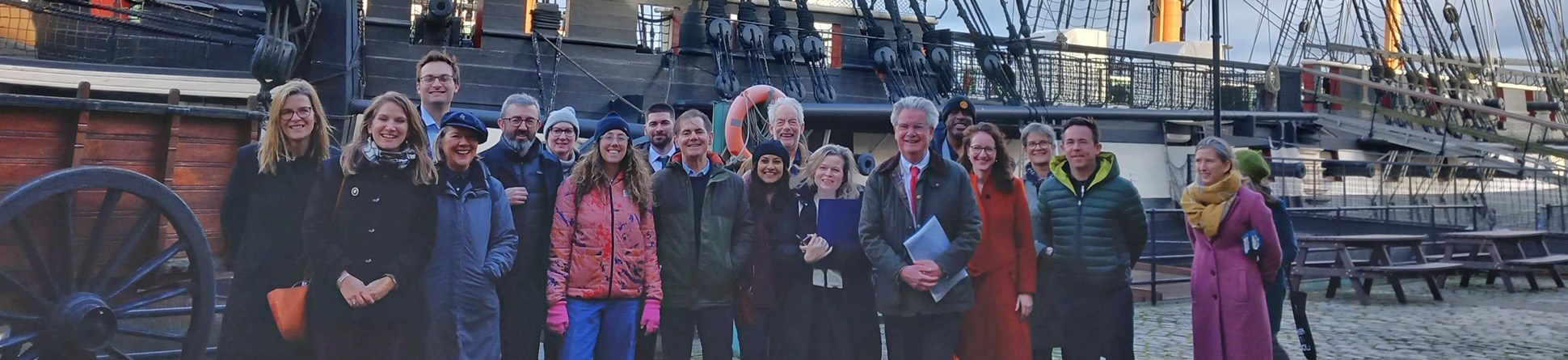 A group of people standing in front of an old sailing boat. 