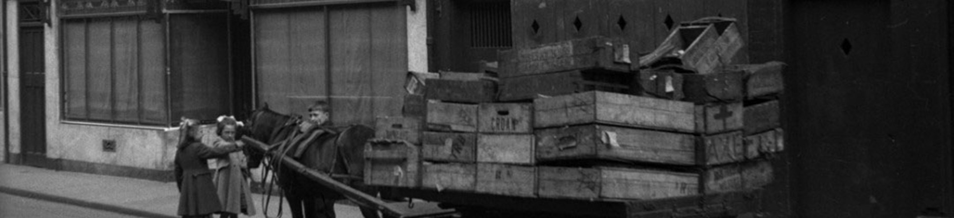 A black and white photograph of a street with brick buildings and old shop fronts with a horse and cart into front of them.
