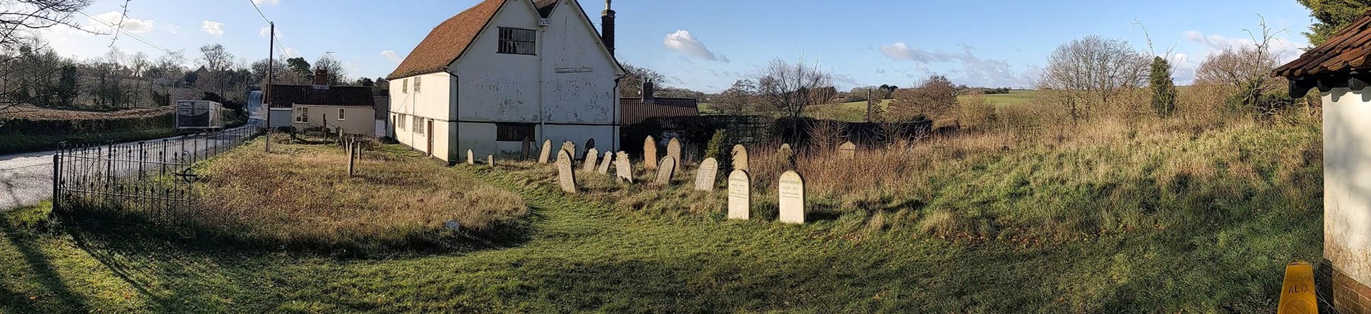 The photograph shows a white building with double roof in brown tiling, with gravestones dotted around and a scrub landscape