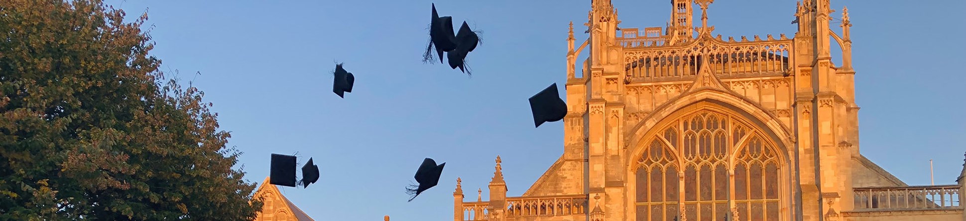 A group wearing graduation gowns throw their mortar boards in the air to celebrate. Behind them, light from the setting sun turns Gloucester Cathedral orange against a blue sky.