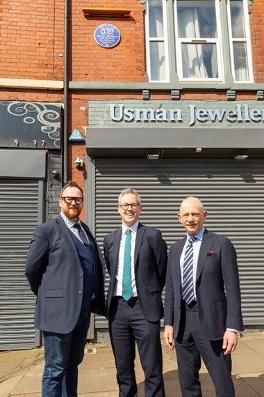 Three men in suits stood in front of a red brick building with grey metal shutters and a blue heritage plaque installed next to a first storey window