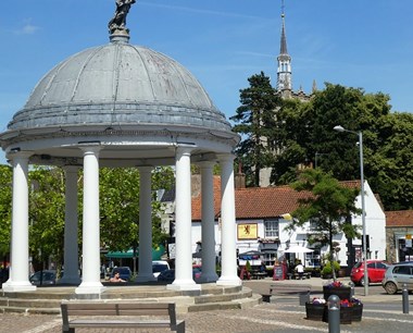 Domed structure resting on seven white pillars. In the background is a two-storey white-painted pub with sign that reads: Red Lion.