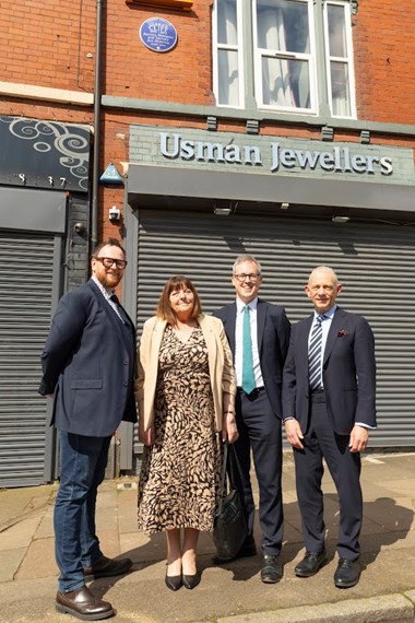 Four people stood in front of a red brick wall building with grey metal shutters and a blue plaque installed next to a first floor window