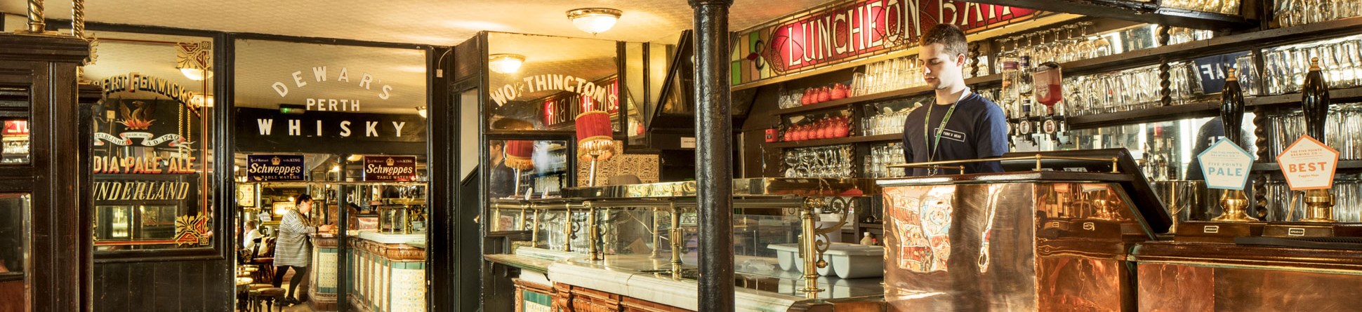 Bar with tiled front and sign behind saying 'LUNCHEON BAR'. On a glass door and mirrored walls there's advertising for various drink brands.