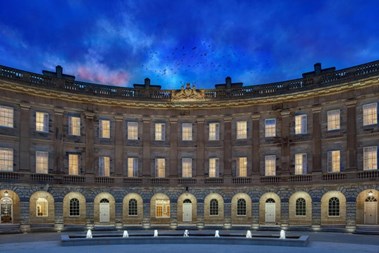 A front-on view of a Georgian-era building underneath a dark blue night sky, with a wide water fountain in the foreground.