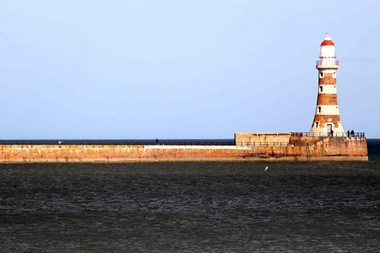 Sea in the foreground. Long pier from left leading to red and white striped lighthouse on the right