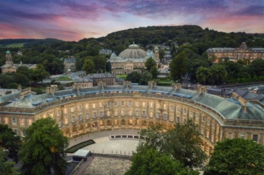 An aerial photograph of a crescent-shaped Georgian-era building lit up at night, with countryside and a sunset sky in the background