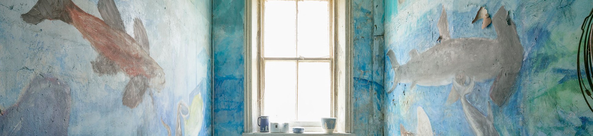 A photograph of a small bathroom with a window frame in the centre and walls painted with a mural of an underwater scene containing fish, a hammerhead shark and other sea life.